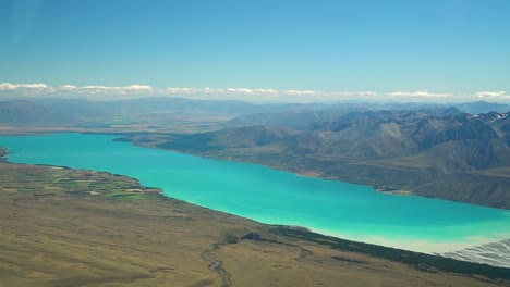 SLOWMO---Aerial-view-of-beautiful-blue-glacier-Lake-Tekapo,-South-Island,-New-Zealand-from-airplane-scenic-flight-and-braided-rivers-with-rocky-snow-capped-mountains-in-background