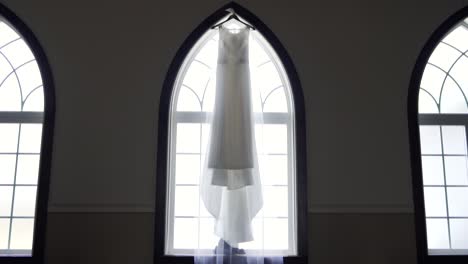 Wide-vista-of-a-bride's-beautiful-wedding-dress-hanging-from-a-large-window-sill-in-an-old-church-building---Slow-push-in-slider-shot