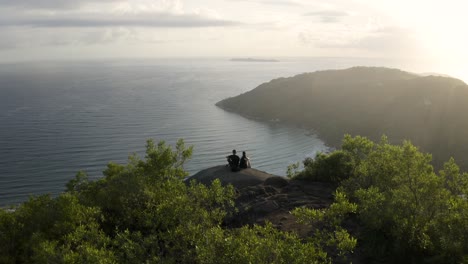 Beautiful-cinematic-drone-establishing-shot-approaching-a-couple-seated-in-a-hiking-peak-spot-of-a-tropical-forest-mountain-revealing-a-beautiful-beach-ocean-scenery-in-the-background