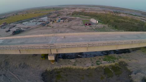 Vista-Aérea-Del-Puente-Construido-Sobre-El-Río-Con-Camiones-Pesados-Y-Maquinaria,-Granjas-En-El-Río