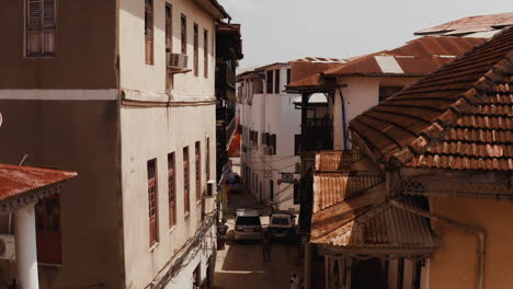 Rise-up-shot-of-an-old-narrow-street-of-Stone-Town,-with-driving-cars,-walking-tourist