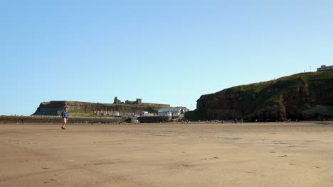 4K-static-camera-showing-man-walking-alone-across-quiet-beach-with-Whitby-Abbey-on-hill-in-background
