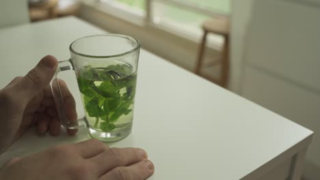Hands-on-table-with-herbal-tea-in-a-glass-in-cozy-home-with-garden