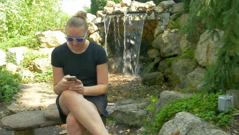 Woman-Laughs,-Smiles,-And-Shakes-Her-Head-While-Looking-At-Her-Smart-Phone-Next-To-A-Small-Waterfall-In-The-Park