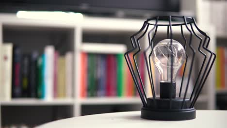 Close-up-on-a-stylish-lamp-that-stands-on-a-white-table-in-the-background-are-books-as-a-blurred-background