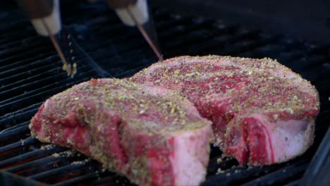 Eine-Fleischzange-Spielt-Mit-Einem-Rohen-Rib-Eye-Steak-Auf-Einem-Grill-Und-Drückt-Darauf-Und-Ein-Weiteres-Steak