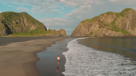 SLOWMO---Aerial-drone-shot-of-young-man-walking-on-black-sand-vulanic-Whatipu-Beach-by-ocean-towards-incredible-rock-formations