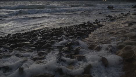 Slow-motion-of-waves-rolling-in-over-rocks-on-foreshore-causing-ocean-foam-on-windy-dusk-evening
