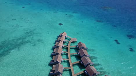 Las-Cabañas-De-Bungalows-Sobre-El-Agua-En-La-Playa-De-Maldivas-Dan-Belleza-A-Las-Aguas-Poco-Profundas-De-Color-Azul-Claro-Y-Al-Agua-Azul-Profunda---Toma-Aérea