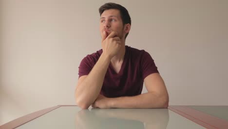 Young-man-facing-camera-thinking-and-wondering-about-something-with-minimalistic-background,-white-wall-and-empty-table,-still-shot