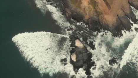 Aerial-descending-of-rocky-formations-while-the-waves-crash-against-it-on-a-cloudy-day-in-Pichilemu,-Chile-4K
