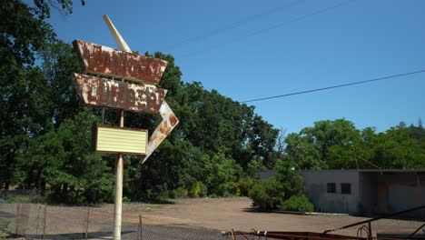 Rotating-left-pan-shot-of-a-rusty-retro-store-sign-out-front-of-an-abandoned-lumber-yard-on-a-dirt-road-with-mountains-in-the-background-on-a-bright-sunny-day