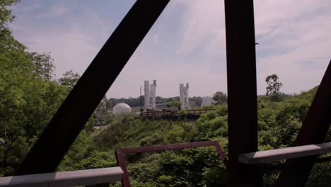 A-metal-trestle-bridge-built-over-a-nature-preserve-with-industrial-silos-in-the-background