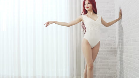 Schöne-Asiatische-Ballerina-Frau-Im-Weißen-Bodysuit-Mit-Langen-Roten-Haaren,-Die-Ballettschuhe-Trägt-Und-Im-Weißen-Raum-Mit-Wand--Und-Vorhanghintergrund-Posiert