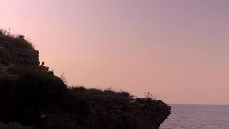 Zwei-Silhouettierte-Menschen-Sitzen-Auf-Einer-Klippe-Vor-Dem-Hintergrundbeleuchteten-Rosafarbenen-Sonnenuntergangshimmel-An-Der-Amalfiküste
