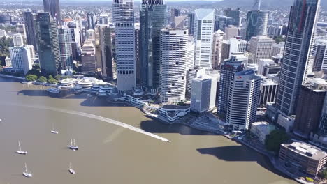 Aerial-tilt-shot-of-tall-city-buildings,-jet-ski-and-sail-boats-along-river-Brisbane,-Queensland
