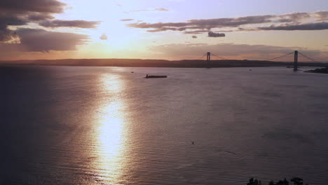 drone-camera-pan-right-over-Coney-Island-Creek-viewing-a-dark-golden,-cloudy-sunset-while-revealing-the-Verrazano-Bridge