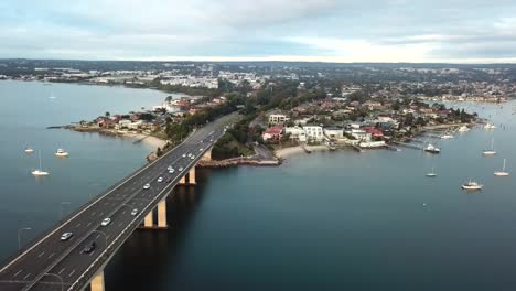 Aerial:-Drone-shot-tracking-cars-driving-across-a-bridge-towards-a-waterfront-suburb-in-Sydney,-Australia