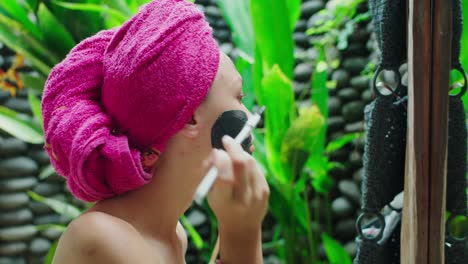 Young-woman-with-towel-on-head-applying-clay-mask-on-her-face-after-shower-in-tropical-Bali-style-bathroom