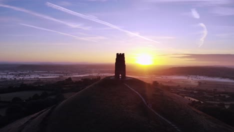 Aerial-pan-of-Glastonbury-Tor-at-sunrise,-with-a-deep-morning-blue-sky