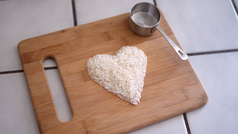 Weißes-Reiskorn-In-Einem-Herzen-Mit-Kochutensilien-Und-Einem-Messbecher-In-Einer-Küchenrutsche-Nach-Rechts