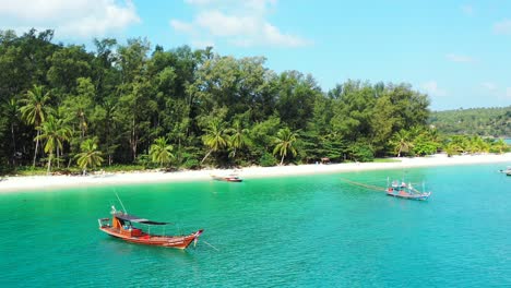 Peaceful-exotic-beach-with-palm-trees-over-white-sand,-calm-turquoise-lagoon-with-anchored-boats-for-touristic-trips-in-Thailand
