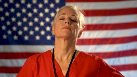 Medium-tight-portrait-of-a-healthcare-nurse-taking-glasses-off-and-looking-concerned-and-sympathetic-with-an-out-of-focus-American-flag