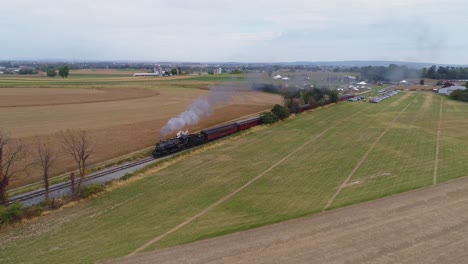 Aerial-angled-view-of-a-restored-steam-engine-blowing-steam-and-smoke-while-pulling-antique-passenger-cars-with-view-of-farmlands-on-a-sunny-day