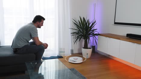 Man-sits-on-a-couch-in-the-living-room-and-activates-his-vacuum-robot-cleaner-via-smartphone-voice-control