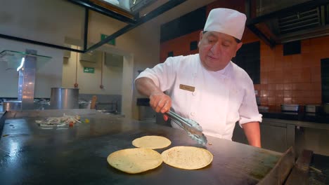 Older-Adult-Male-Mexican-chef-flips-tortillas-on-a-grill,-while-vegetables-cook-in-preparation-for-fajitas