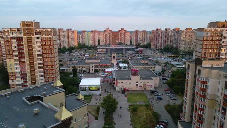 Aerial-view-over-a-street,-towards-stores-and-markets,-in-middle-of-sunlit-apartment-buildings,-in-Troieshchyna-soviet-district,-during-sunset,-in-Kiev-city,-Ukraine---dolly,-drone-shot