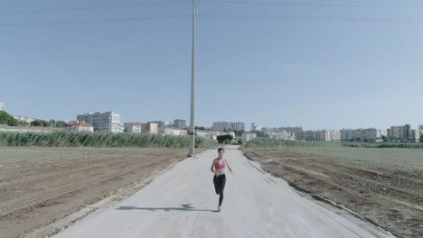 Wide-Drone-shot-of-Young-Woman-running-in-Slow-Motion-towards-the-camera-in-urban-park-dirt-road