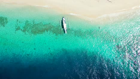 Anchored-boat-floating-on-clear-water-of-turquoise-lagoon-which-flows-on-white-sand-of-exotic-beach,-copy-space-with-colorful-sea-texture