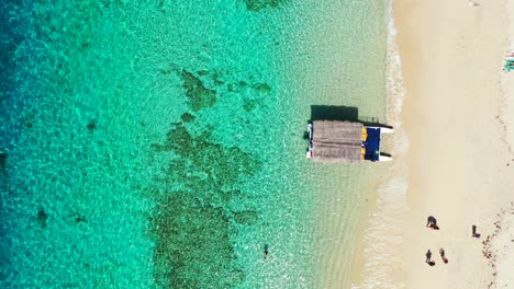 Traditional-boat-with-straw-roof-anchoring-on-sandy-beach-washed-by-calm-clear-water-of-blue-turquoise-lagoon-with-corals-in-Philippines