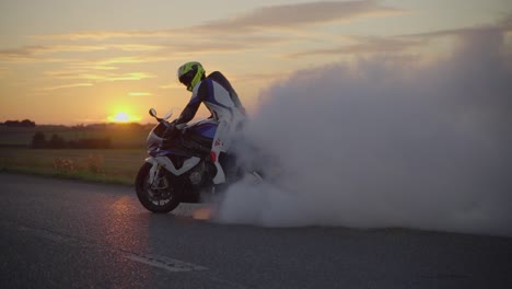 Medium-wide-shot-of-a-motorbiker-wearing-full-body-suit-and-helmet-performing-a-burnout-with-a-white-superbike-during-sunset