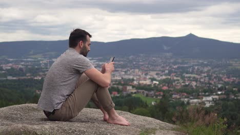 Kaukasischer-Mann-Benutzt-Draußen-Sein-Mobiltelefon,-Tippt-Und-Wischt,-Sitzt-Auf-Einem-Felsen-Und-Hat-Eine-Atemberaubende-Aussicht-Im-Hintergrund