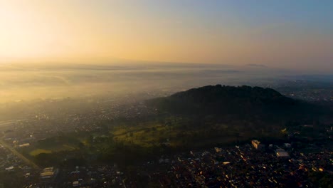 Mount-Tidar-on-picturesque-sunrise-and-Magelang-city-covered-in-fog,-aerial-view