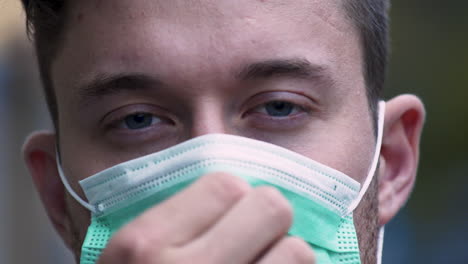 A-young-man-with-bright-blue-eyes-and-a-stubble-puts-on-a-green-protective-Covid-19-facemask-in-public,-adjusting-it-on-his-face,-while-looking-straight-into-the-camera,-static-close-up-4k-shot