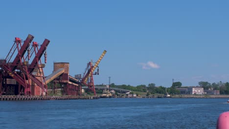 Port-cranes-and-harbor-warehouses-in-sunny-calm-summer-day-at-Port-of-Ventspils,-Venta-river-in-front-of-frame,-wide-shot-from-a-distance