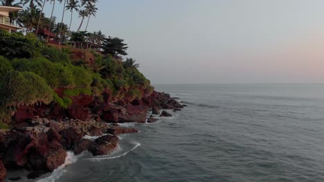 Forward-moving-aerial-along-the-tall-dense-green-cliffs-of-Varkala-Beach,-Kerala,-India-showing-low-tides-hitting-the-rocks