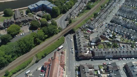 Aerial-of-train-line-and-residential-streets-in-urban-england-on-a-sunny-day,-river-and-community-facilities-also-visible