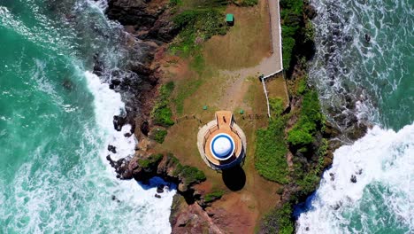 Overhead-view-of-structure-at-tip-of-rocky-seaside-peninsula-with-green-ocean-waves-crashing-onshore,-Puerto-Plata,-Dominican-Republic,-above-aerial-rising
