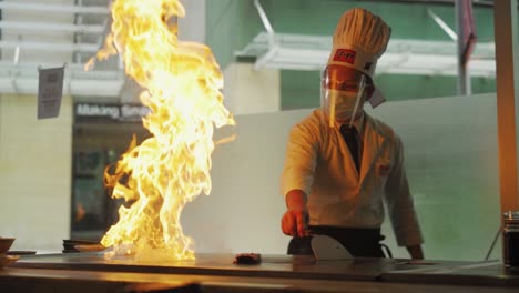 Teppanyaki-Show---Chef-With-Toque-Cooking-On-Iron-Griddle-With-Blazing-Fire-In-Asian-Restaurant