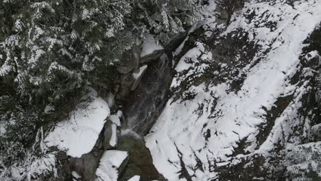Schnell-Fließendes-Wasser-über-Einen-Wasserfall,-Während-Weiße-Schneeflocken-Zwischen-Den-Kiefern-Und-Felsen-In-Einer-Winterszene-Herabwirbeln