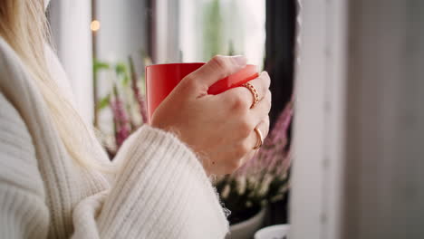 closeup-of-hands-of-a-young-woman-holding-a-big-red-mug-of-coffee,-standing-at-the-window