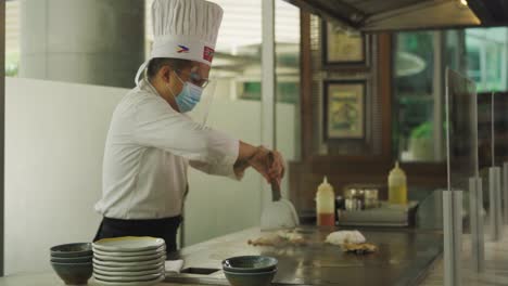 Chef-Wearing-Mask-And-Face-Shield-Cooking-Fried-Rice-On-Teppanyaki-Grill-In-An-Asian-Restaurant