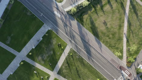 Car-drives-on-quiet-road-next-to-green-park-in-sun,-aerial-drone-shot