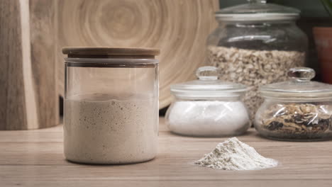 Sourdough-Starter-Yeast-Rising-In-A-Glass-Jar-With-Heap-Of-Flour-On-The-Side---Fermentation---zoom-in-shot,-time-lapse