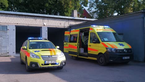 Two-yellow-paramedic-ambulance-vehicles-parked-outside-the-white-brick-garage,-blue-flashing-warning-lights,-sunny-day,-handheld-wide-shot