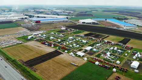 Drone-Shot-with-Ascending-Top-View-of-Agricultural-Expo-Fair-on-Green-Fields-of-Farm-Lands-with-Factories-and-Highway